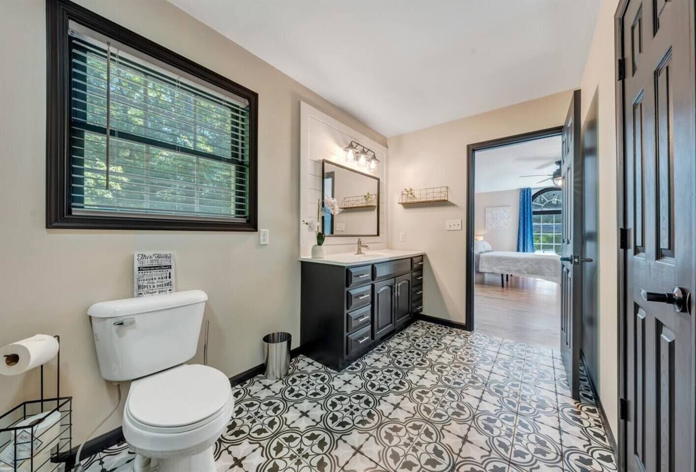 Baño con inodoro, lavamanos y espejo. Hay una ventana con persianas, un cubo de basura y un piso estampado.