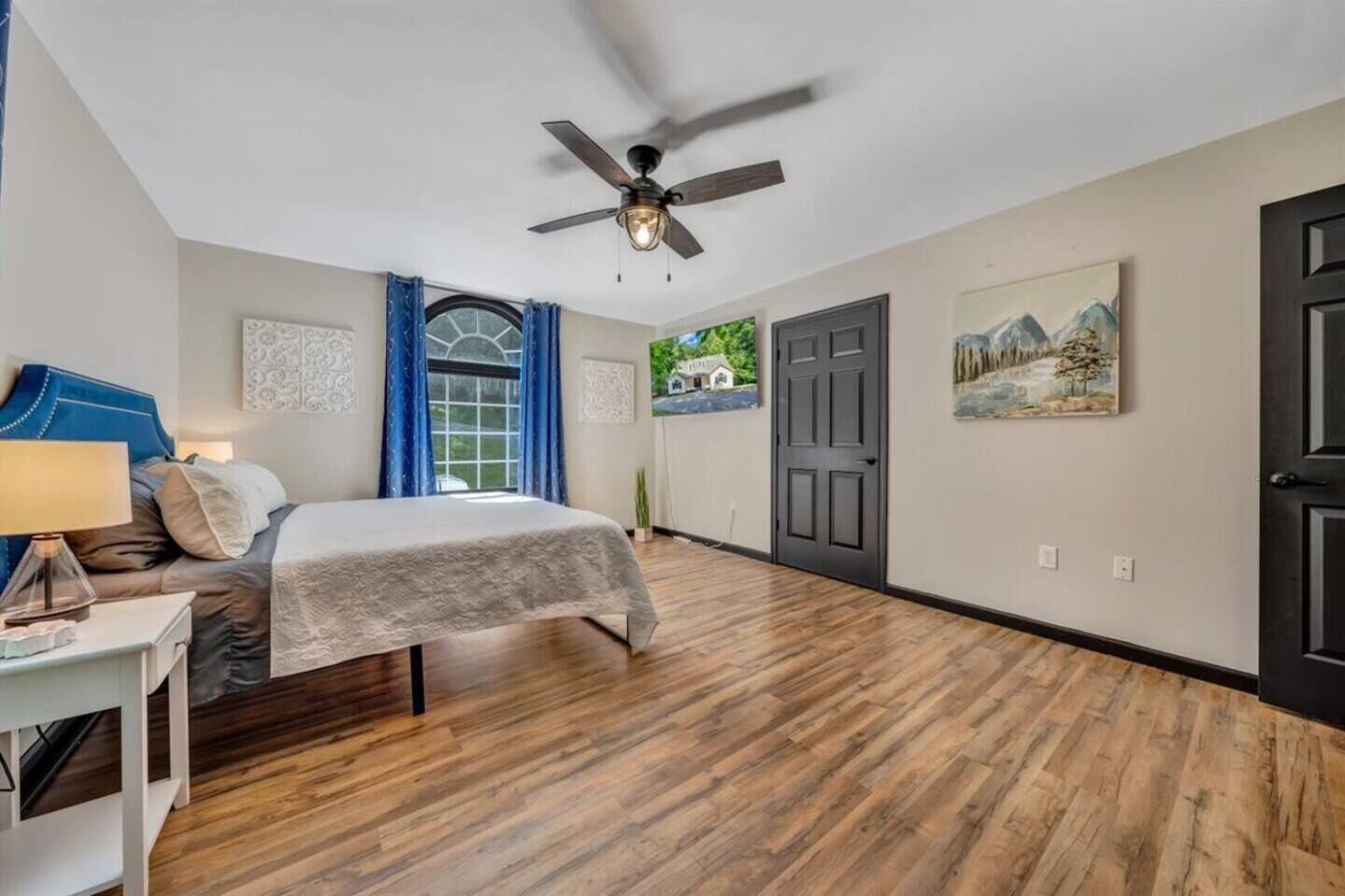 Ein Schlafzimmer mit einem Bett, einem Deckenventilator, einem Fenster mit Vorhängen, einer Tür und einem Bild an der Wand.