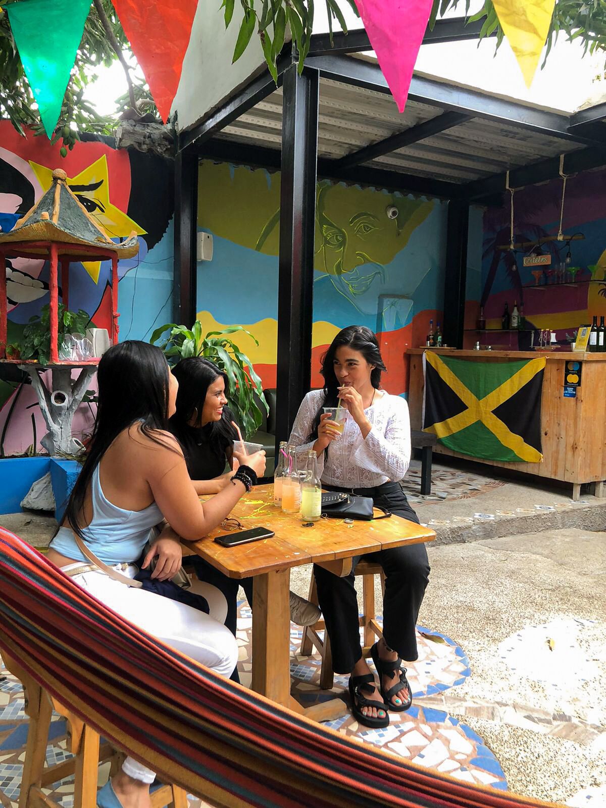 Un grupo de personas sentadas en una mesa al aire libre, con una hamaca cerca, bajo un mural colorido y una estructura de madera.