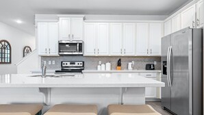 A modern kitchen with white cabinets, a stainless steel refrigerator, a built-in microwave, and a large island with stools. - Townhome with Screened Pool, Kids Bedrooms, Near Attractions (Kissimmee)