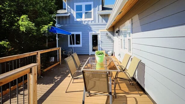 Um deck com mesa e cadeiras, um guarda-sol azul e uma casa ao fundo.