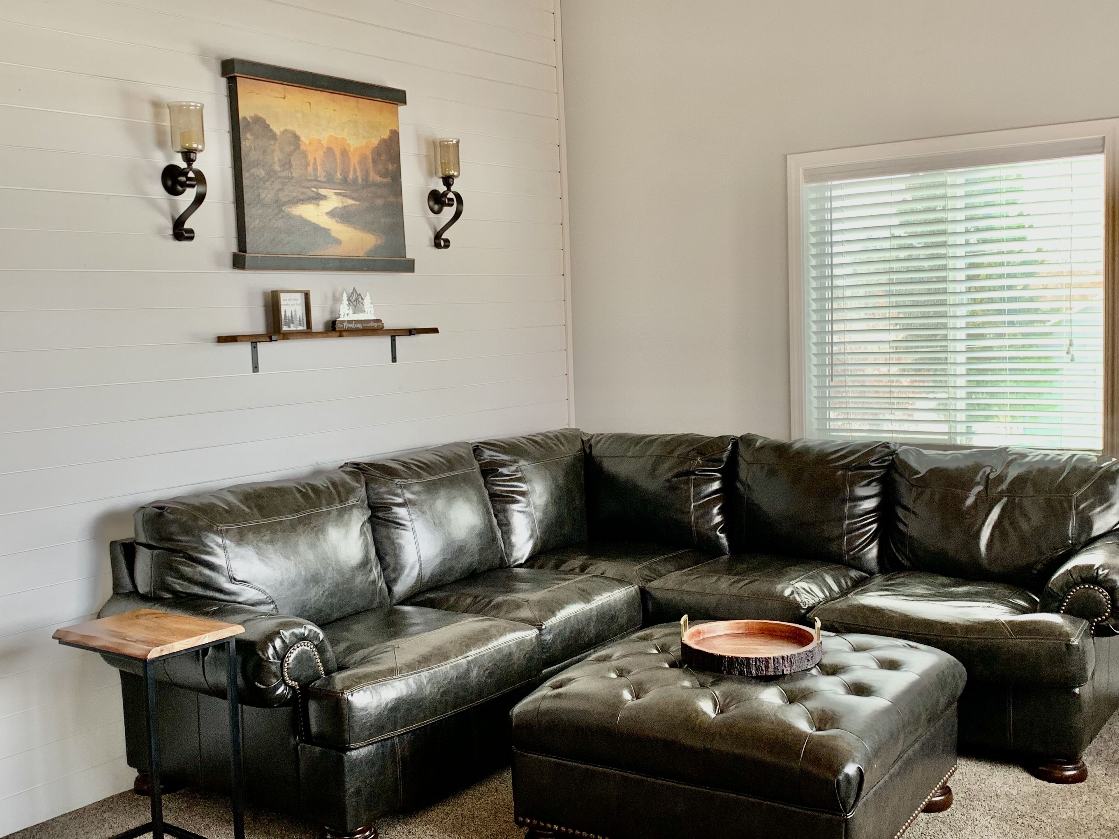 A sectional sofa with a wooden side table, a decorative tray, wall-mounted sconces, and a framed picture on a white paneled wall.