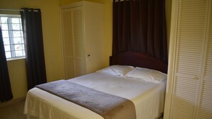 Cama bem arrumada com manta marrom, cabeceira branca e janela com cortinas escuras.