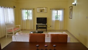Uma sala de estar com sofá, mesa de jantar com banquetas, televisão em móvel e duas janelas com cortinas.