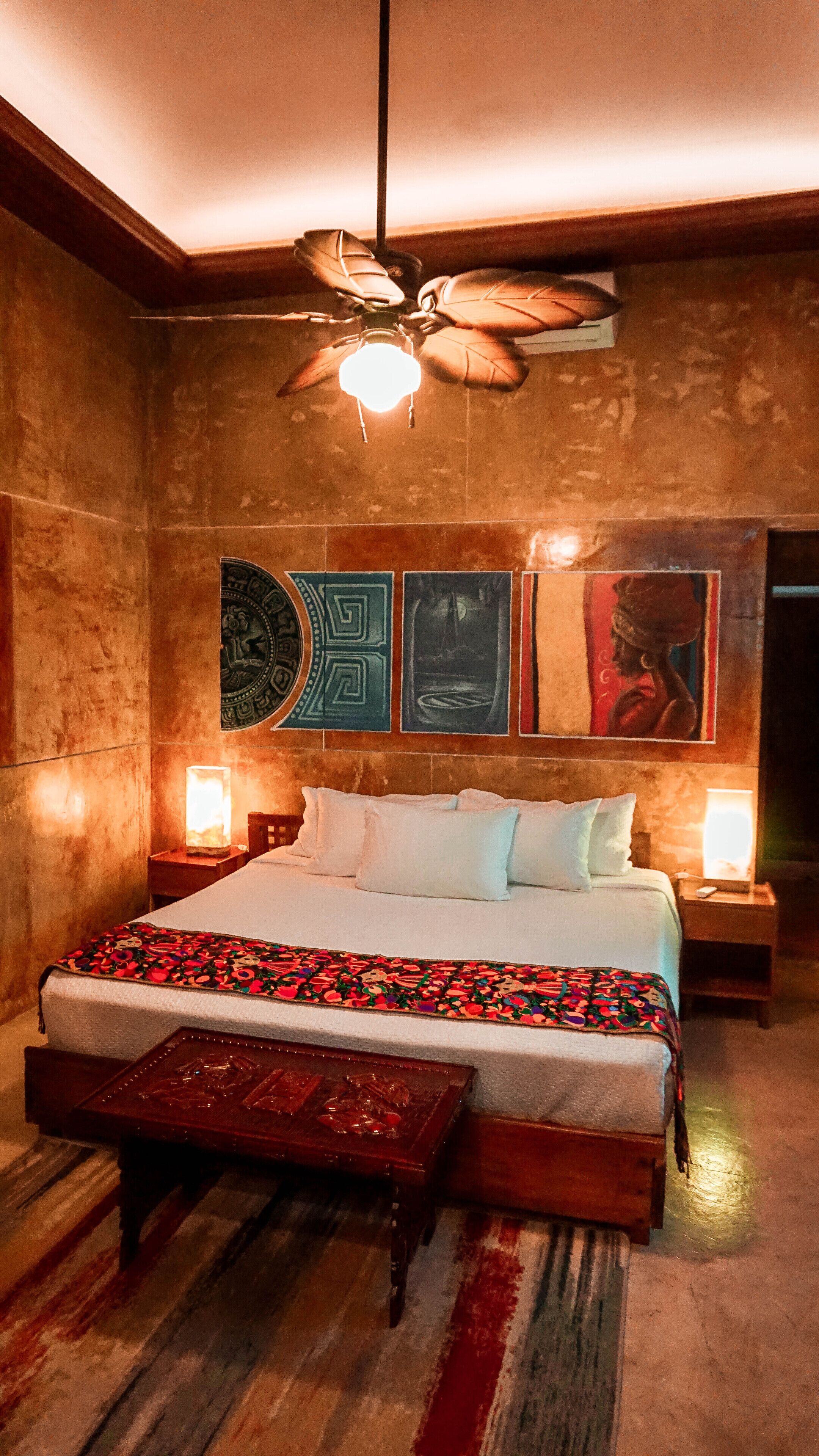 A bedroom with a large bed, a wooden bench, a ceiling fan, and wall art.
