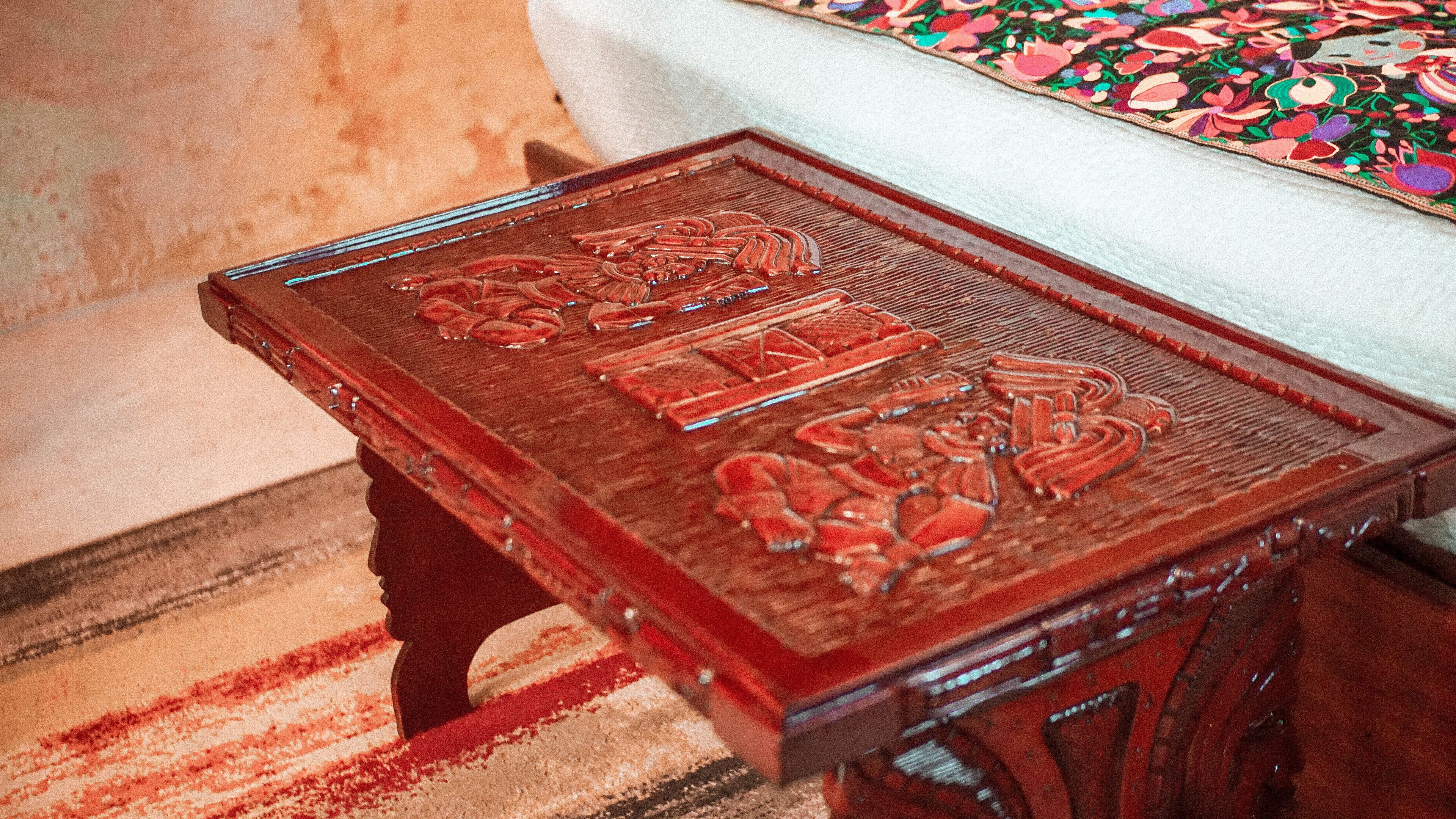 A wooden table with intricate carvings, positioned in a room with a patterned rug and a bed with a floral bedspread.