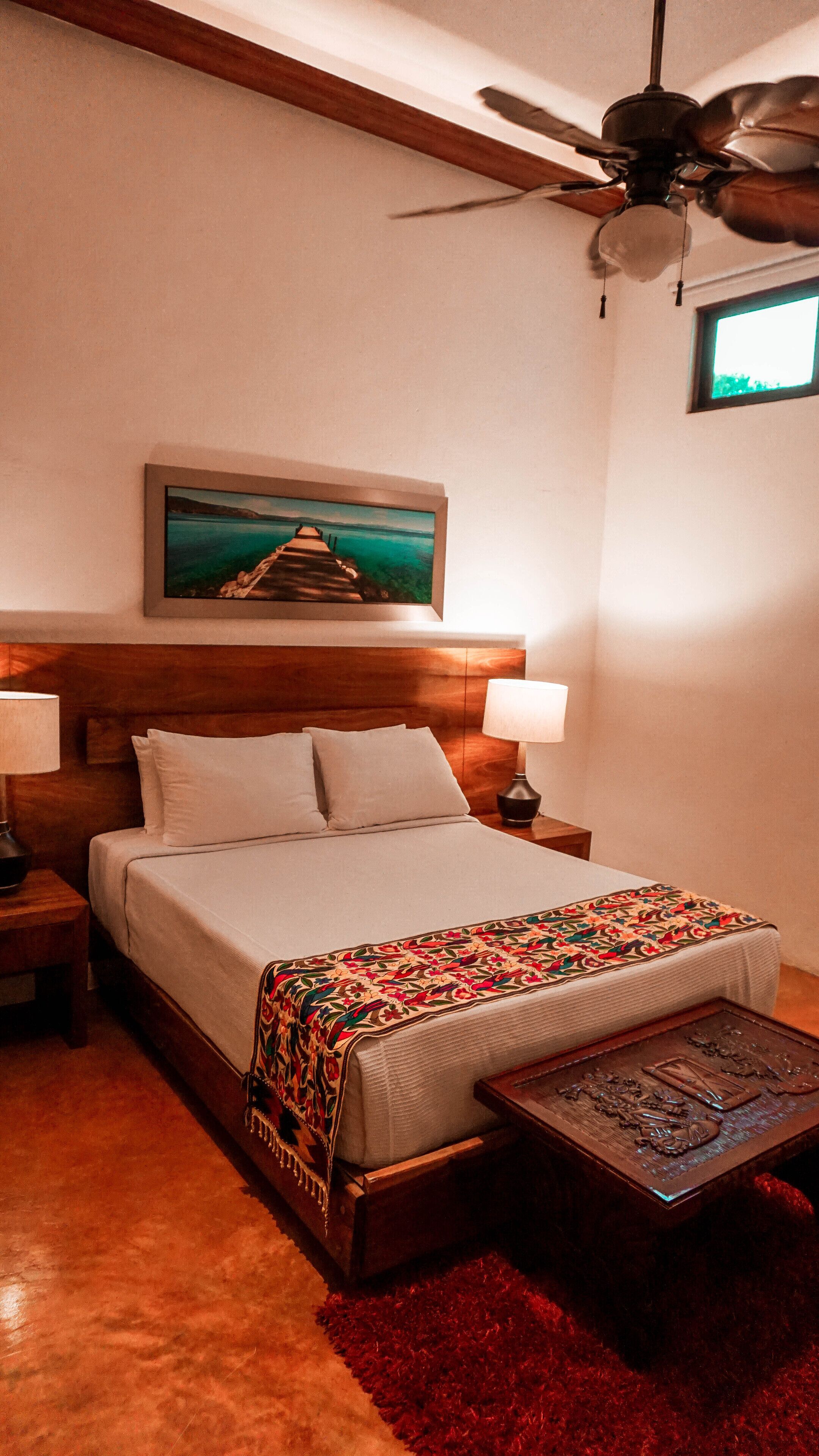 A bedroom with a bed, two bedside tables, a ceiling fan, a painting, and a window.