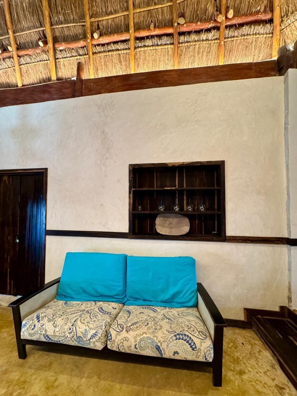 A room with a thatched roof, a sofa with patterned upholstery, a wooden shelf with glassware, and a wooden door.