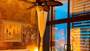 A bedroom with a large bed, a ceiling fan, a painting of sailboats, and a view through a glass door. - ALA Residencias (Chetumal)