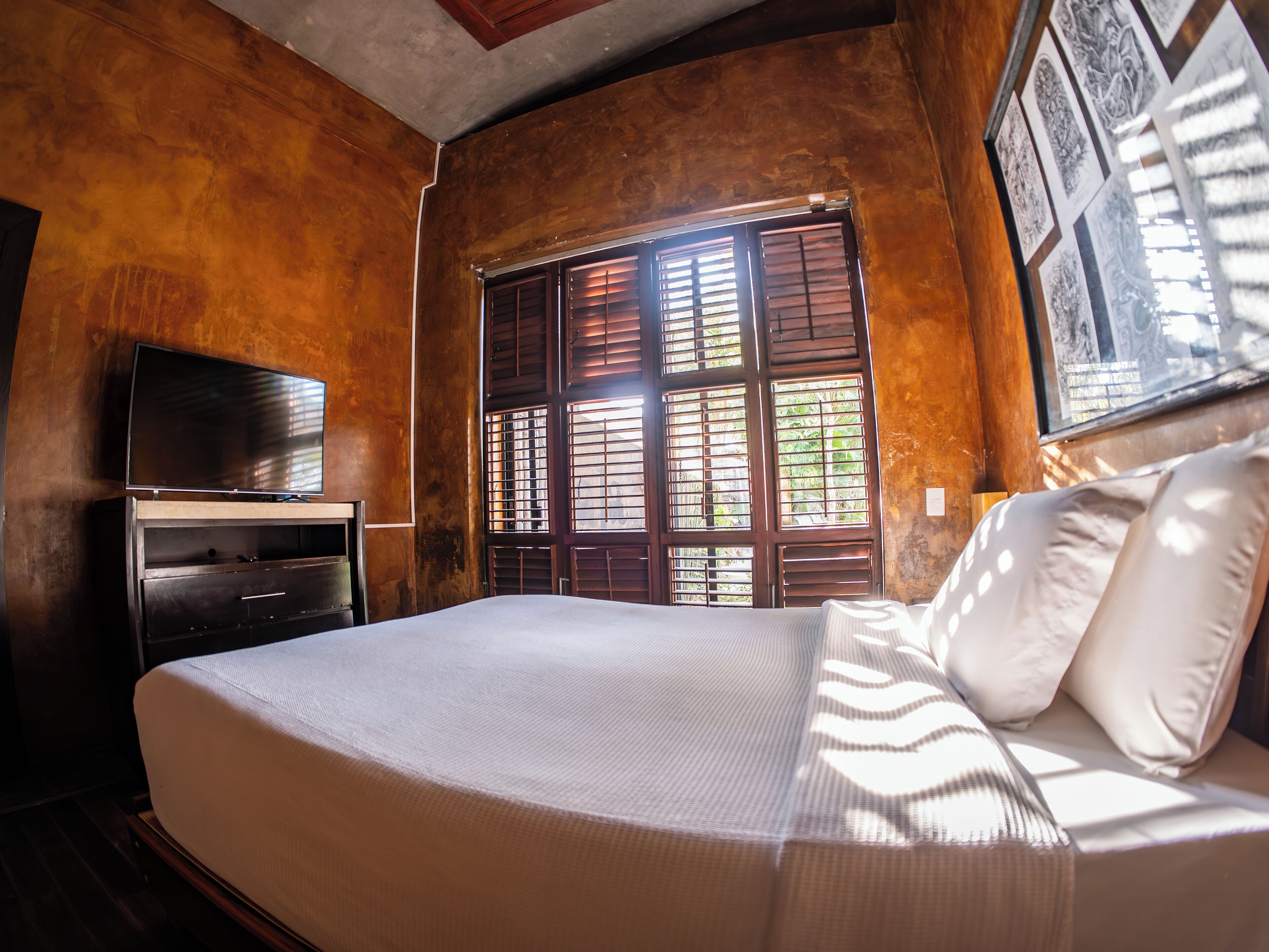 A bedroom with a bed, a TV on a wooden cabinet, and a window with wooden shutters.