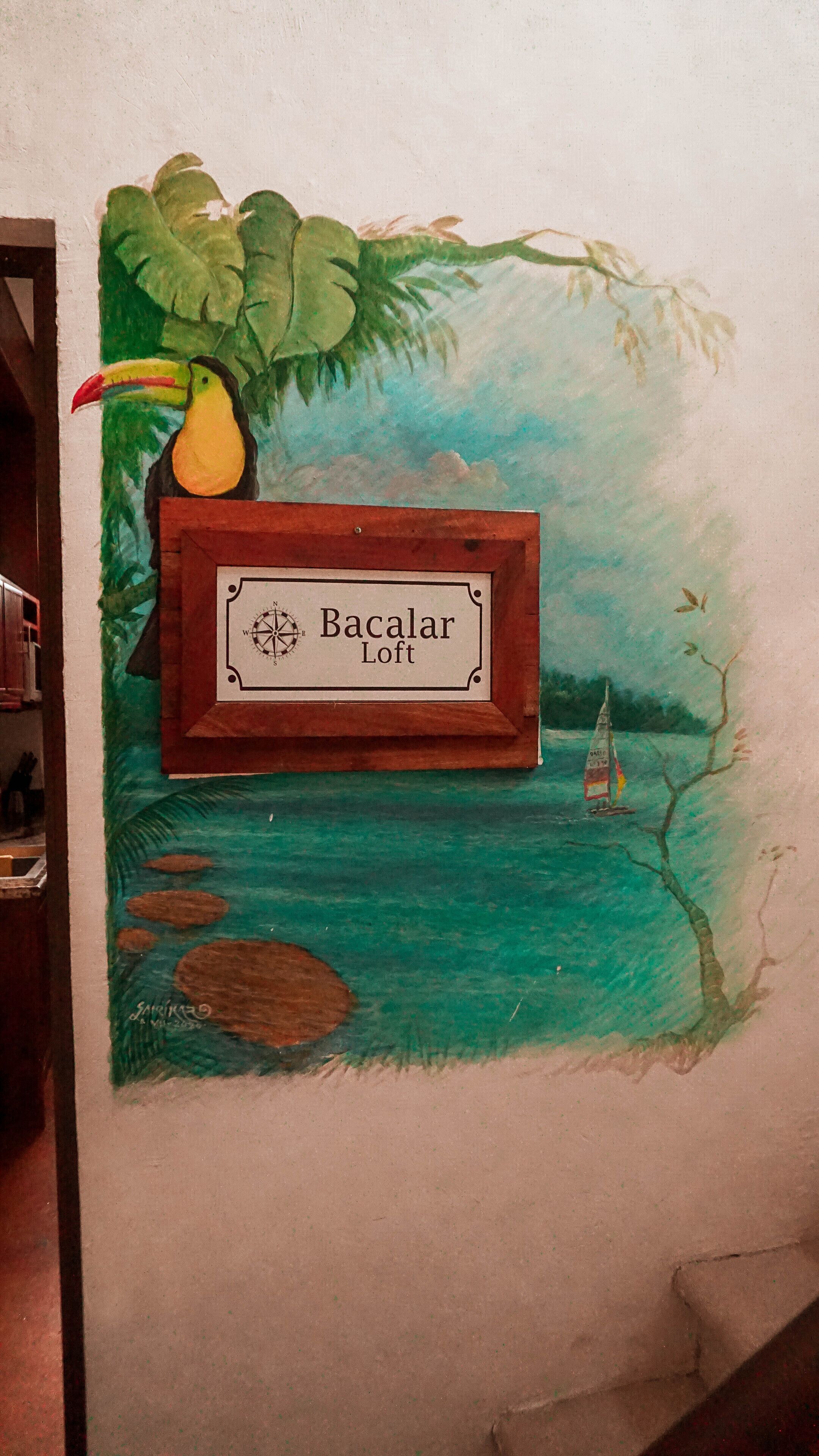 A wall with a painted mural of a toucan, a sailboat, and a compass labeled "Bacalaar Loft".