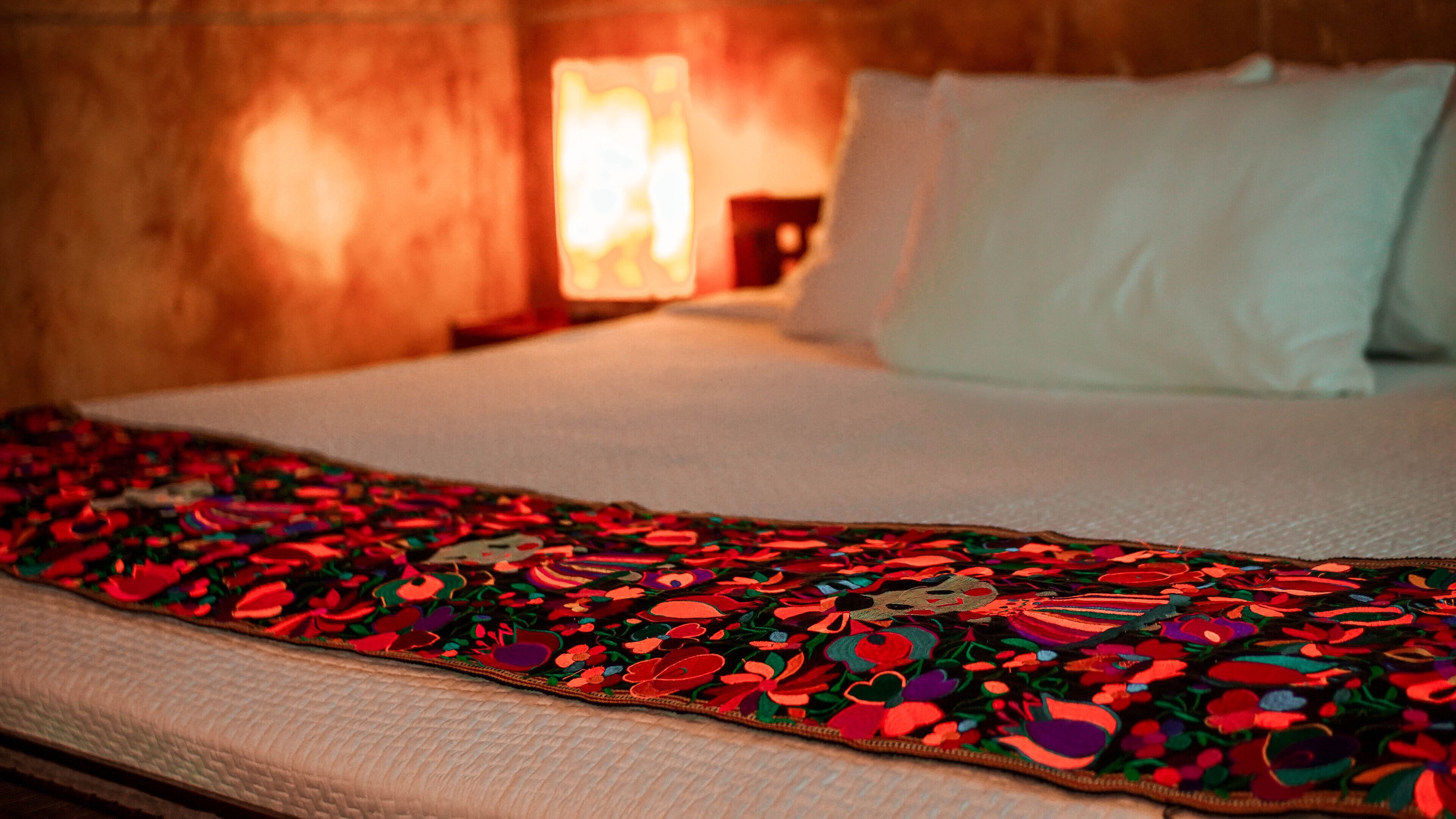 A bed with a colorful blanket, white pillows, and a lamp in the background.