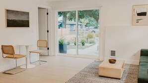 A modern living room with a large sliding glass door leading to a garden, a green sofa, a small white table, and two wicker chairs. - Rose House (Seattle)