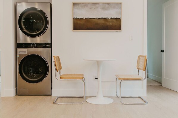 A compact laundry area with a stacked washer and dryer, a small round table, and two wicker chairs. - Rose House (Seattle)