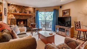 A cozy living room with a stone fireplace, comfortable seating, a television, and a balcony with blue curtains.