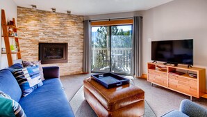 A modern living room with a stone wall, a fireplace, a large TV, and a balcony with a view of trees. - Westwind 302 2BD 2BA Plat. Condo (Vail)
