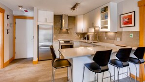 A modern kitchen with white cabinets, stainless steel appliances, and a central island with bar stools. - Westwind 302 2BD 2BA Plat. Condo (Vail)