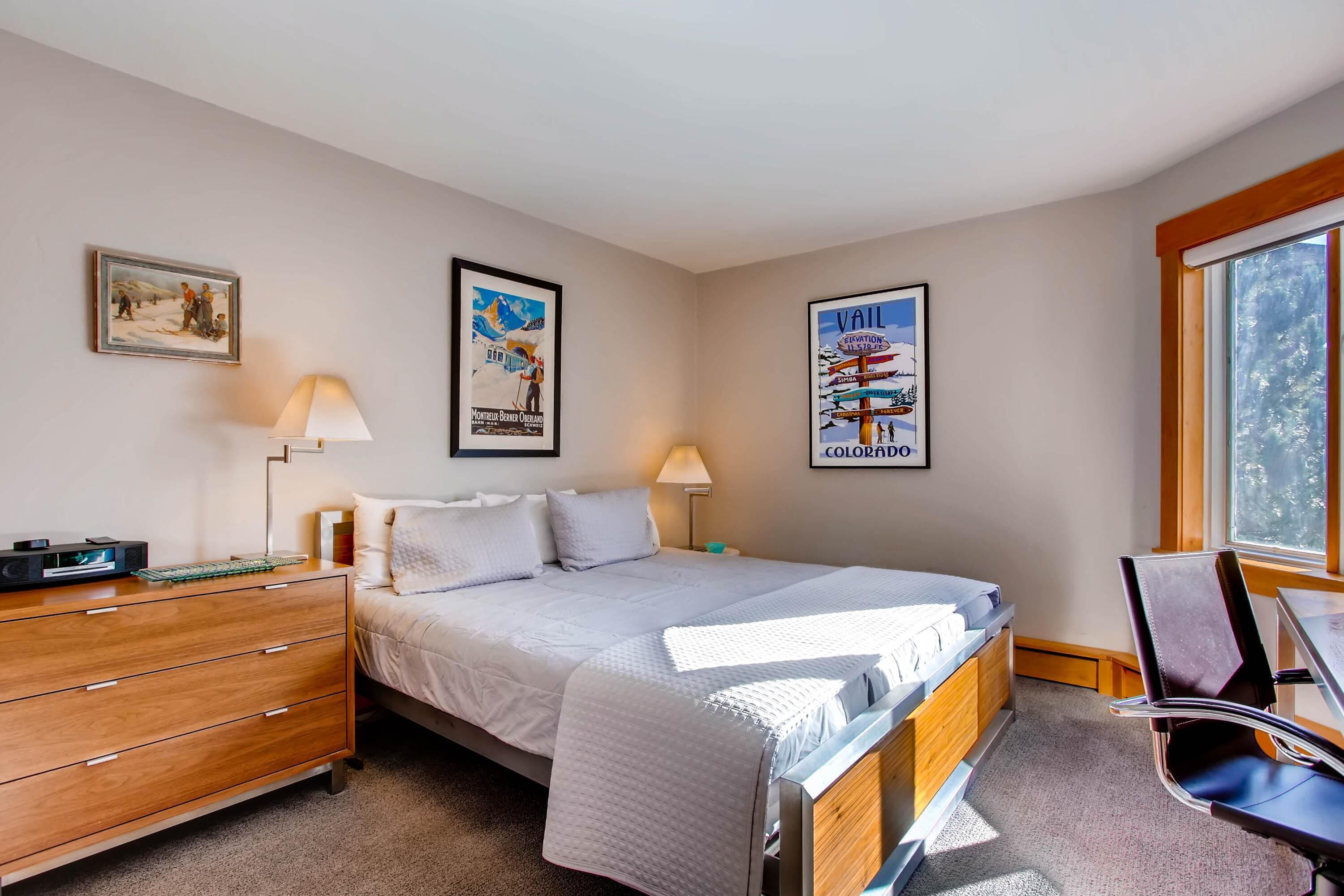 Ein Schlafzimmer mit Bett, Nachttischen, Schreibtisch und Stuhl. An den Wänden hängen Poster und durch das Fenster ist ein Blick auf Bäume zu sehen.