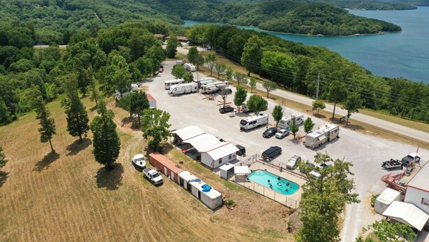 Parque para vehículos recreativos con varias caravanas, una piscina y una vista panorámica de un lago rodeado de colinas boscosas.