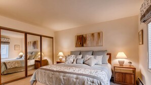 A bedroom with a large mirror, a bed with multiple pillows, bedside tables with lamps, and a large abstract painting on the wall. - Westwind 109 1BD 1.5ba Gold Condo in Lionshead Village (Vail)