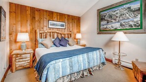 Ein Schlafzimmer mit einem Holzkopfstück, einem Bett mit blauer Bettdecke, zwei Nachttischlampen und einem gerahmten Bild an der Wand.
