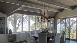 A dining area with a round table, six chairs, and a chandelier. Large windows offer a view of trees and a landscape.