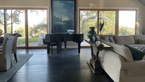 A spacious living room with a grand piano, a large painting, and a view of a garden through large windows.