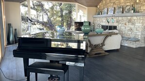 A modern living room with a grand piano, a stone fireplace, and a large window overlooking trees.