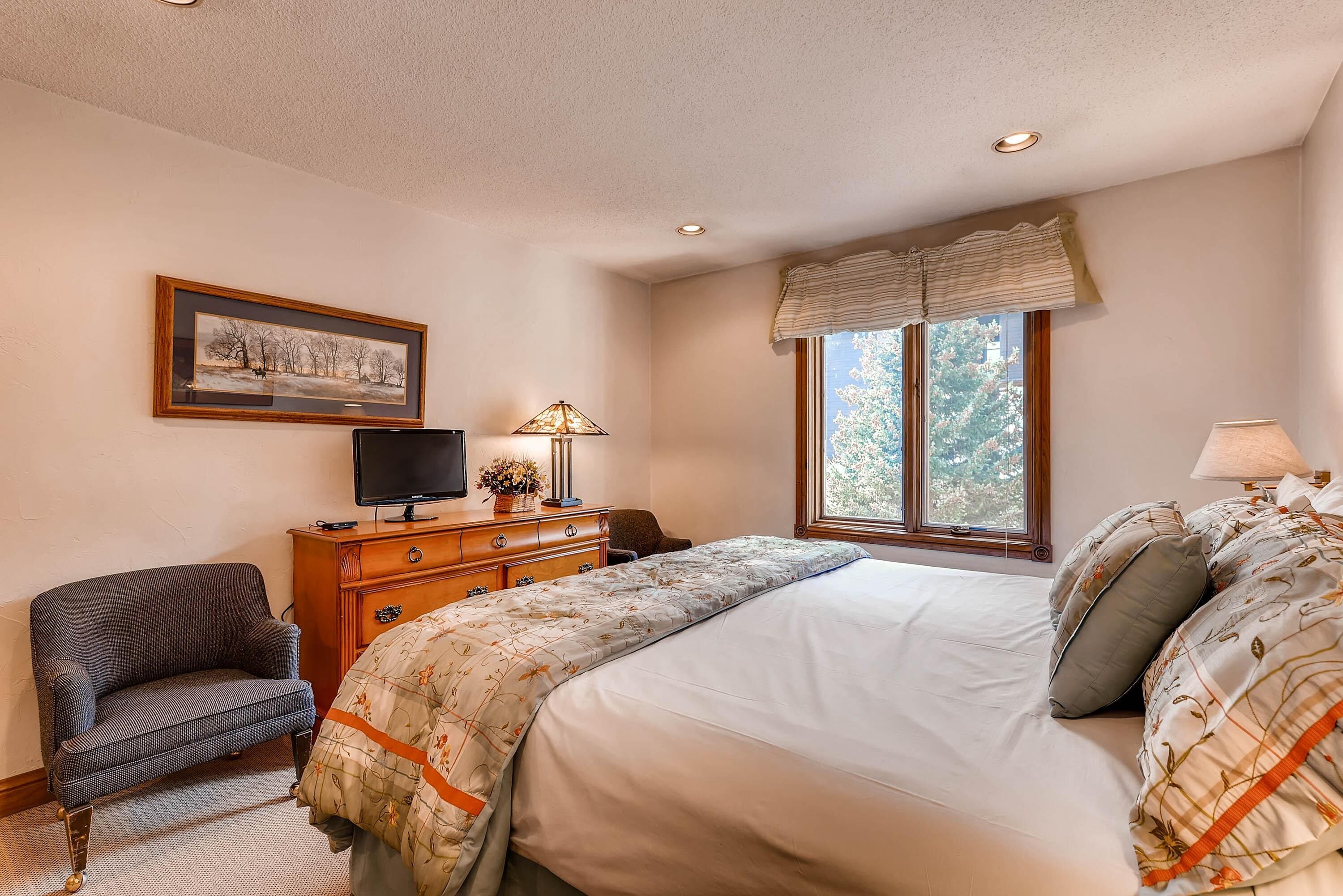 A bedroom with a bed, a chair, a dresser, a television, and a window with curtains.