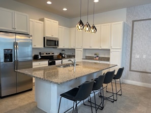 A modern kitchen with white cabinetry, a stainless steel refrigerator, a built-in microwave, a granite countertop, and a bar stool seating area.