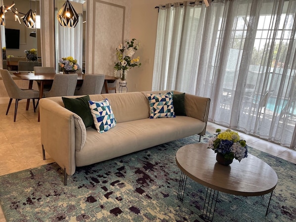 A living room with a beige sofa, patterned cushions, a round coffee table with a vase of flowers, a dining area with a table and chairs, and a large window with sheer curtains.