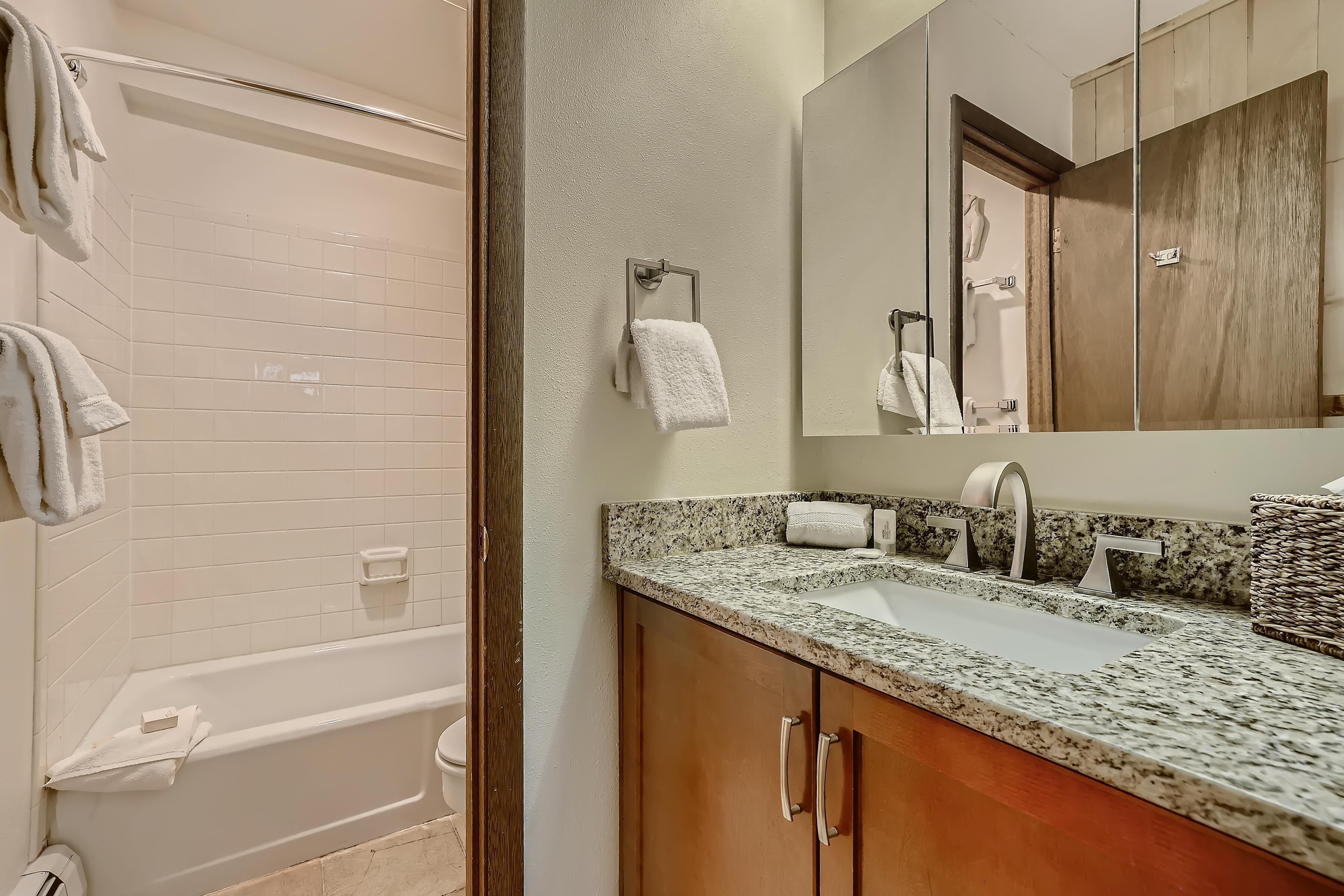 A bathroom with a bathtub, a sink with a granite countertop, wooden cabinets, and a mirror.