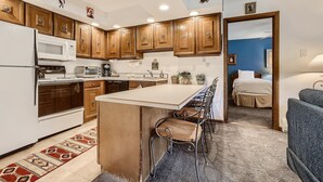 A compact kitchen with wooden cabinets, a refrigerator, microwave, and a dining area with chairs. - Westwind 102 2BD 2BA Diamond Condo in Lionshead Village (Vail)
