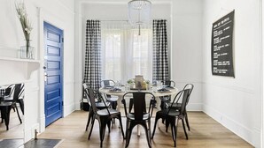 A dining area with a round table, black chairs, and a chandelier. There is a blue door and a wall-mounted sign listing local attractions.