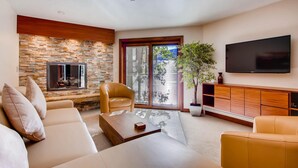 A modern living room with a stone wall, a flat-screen TV, a sofa, a coffee table, and a sliding glass door leading to a balcony. - Westwind 101 2bd, 2BA Diamond Condo in Lionshead Village (Vail)
