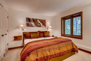 A bedroom with a large bed, a painting above the bed, two bedside lamps, and a window with a view of trees.