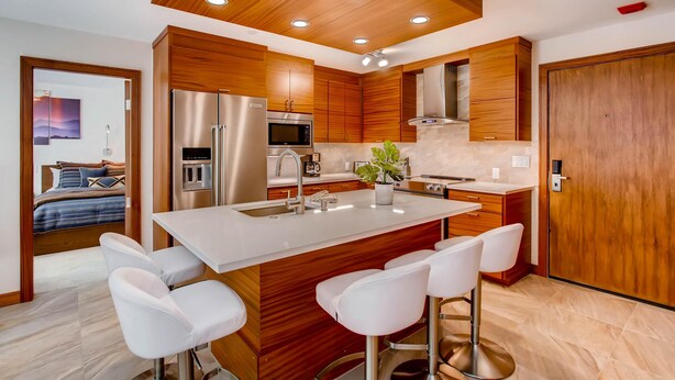 A modern kitchen with wooden cabinets, stainless steel appliances, a central island with bar stools, and a visible bedroom with a bed and artwork. - Westwind 101 2bd, 2BA Diamond Condo in Lionshead Village (Vail)