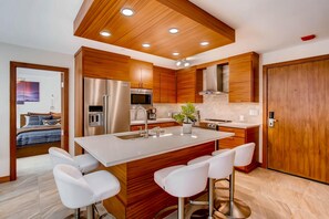 A modern kitchen with wooden cabinets, stainless steel appliances, a central island with bar stools, and a visible bedroom with a bed and artwork.