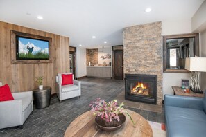 A modern living room with a fireplace, a television mounted on a wooden wall, and a kitchen area in the background.