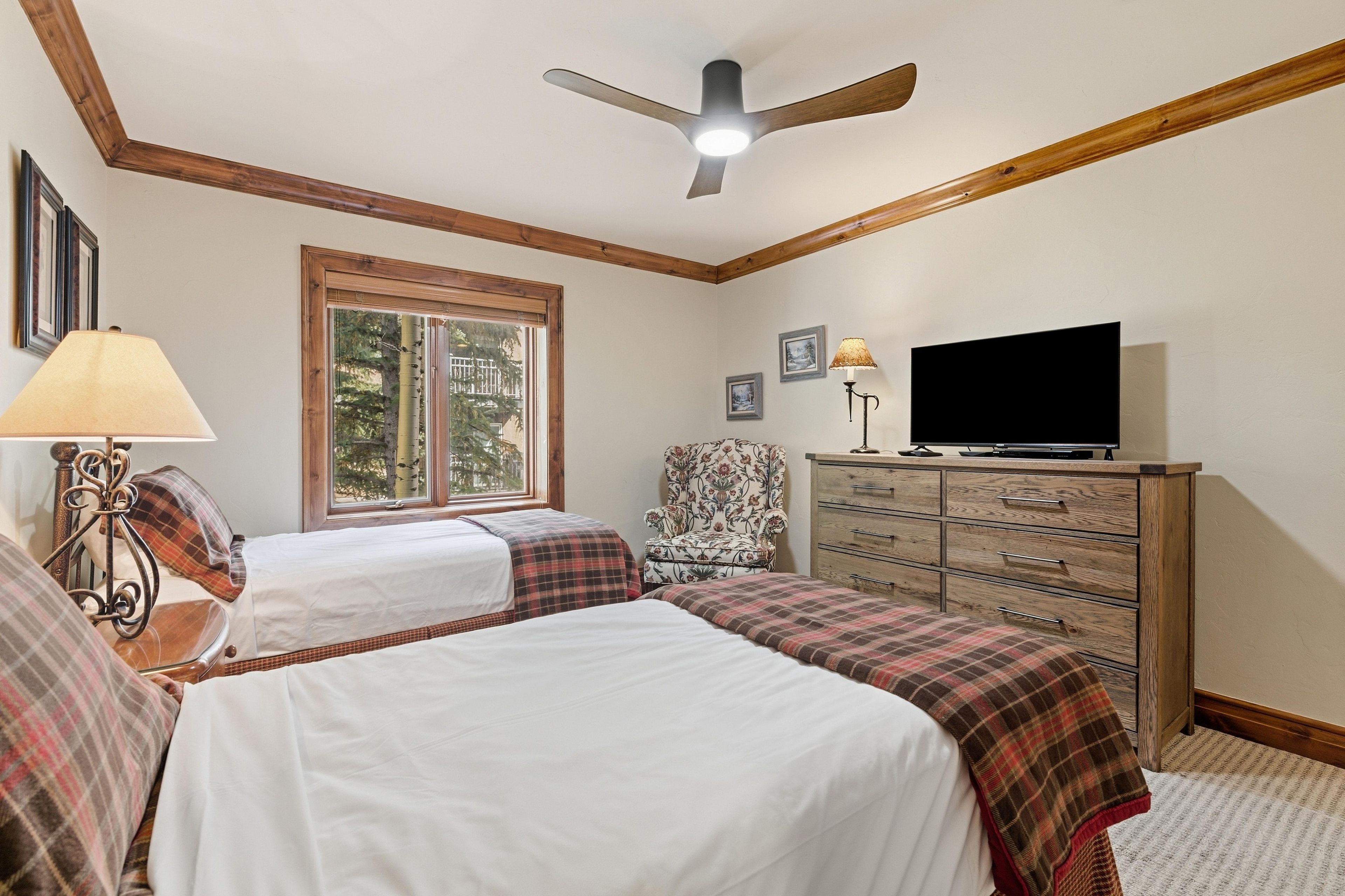 Une chambre avec deux lits, un ventilateur de plafond, un téléviseur posé sur une commode et une fenêtre donnant sur des arbres.