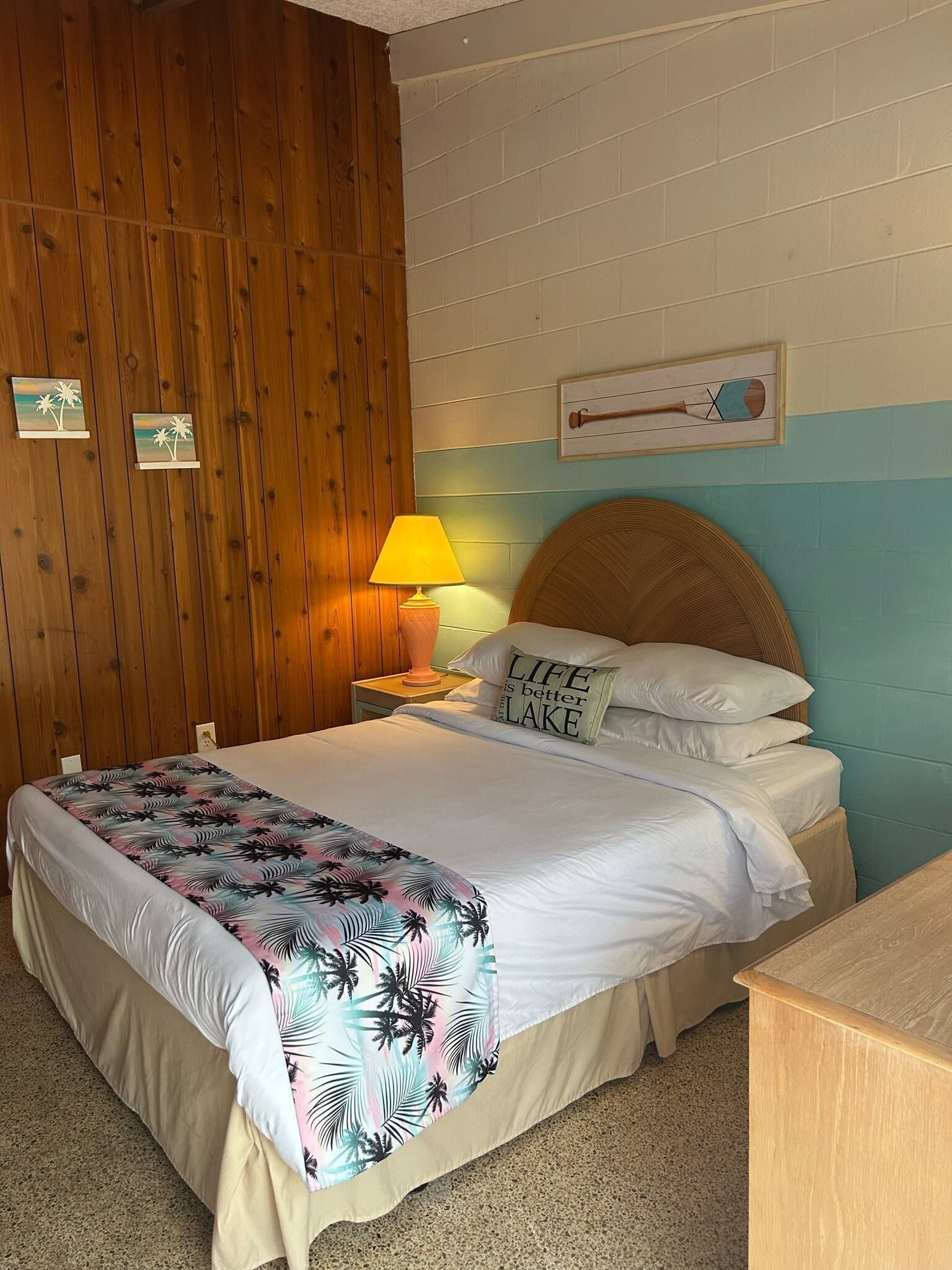 Un letto rifatto con cura, coperto da una trapunta a tema tropicale, una lampada da comodino e un cuscino con la scritta "LIFE is better in LAKE".