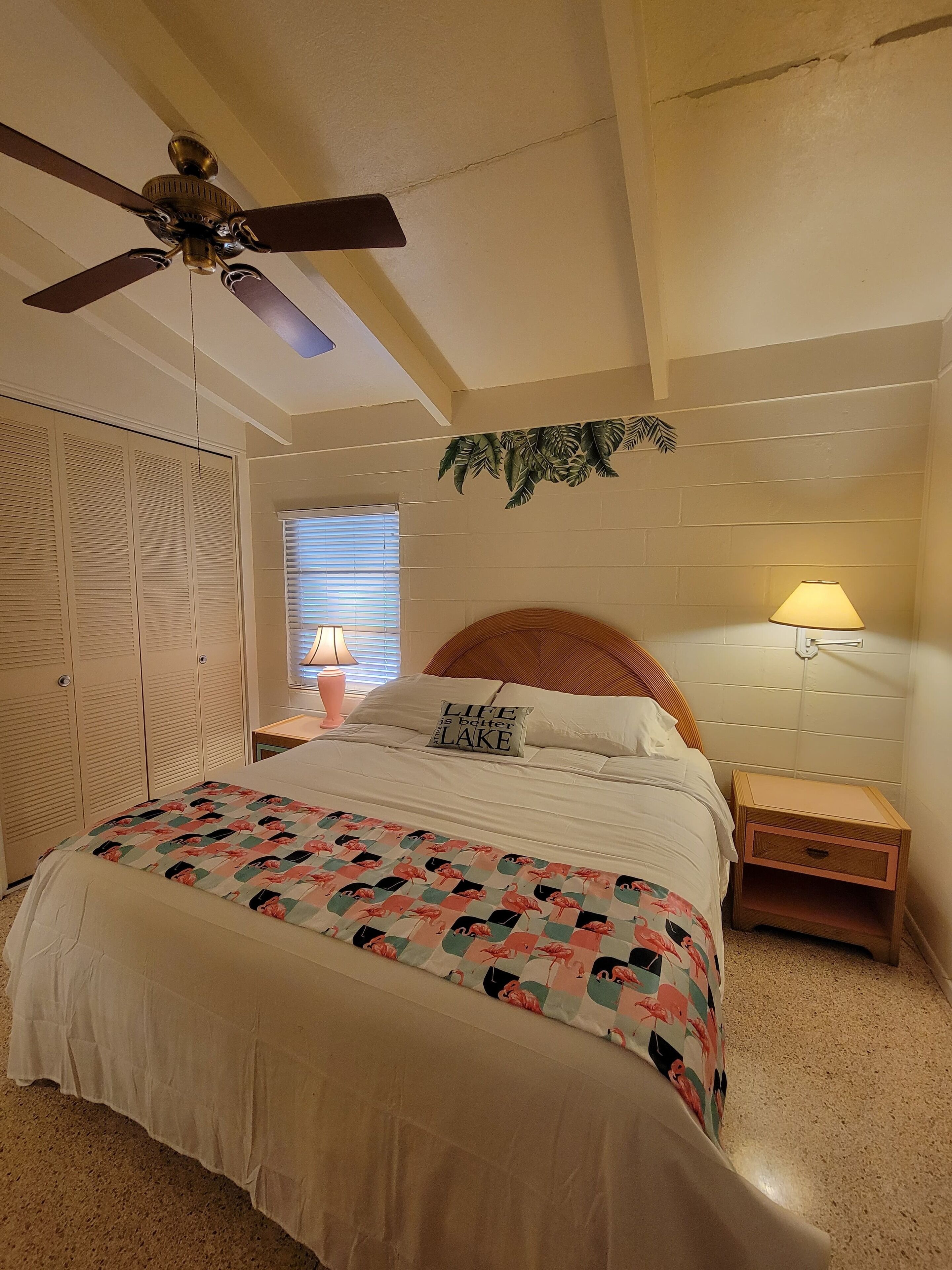 A bedroom with a bed, a ceiling fan, a nightstand with a lamp, a wooden headboard, and a window with blinds.