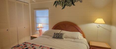 A bedroom with a bed, a ceiling fan, a nightstand with a lamp, a wooden headboard, and a window with blinds.