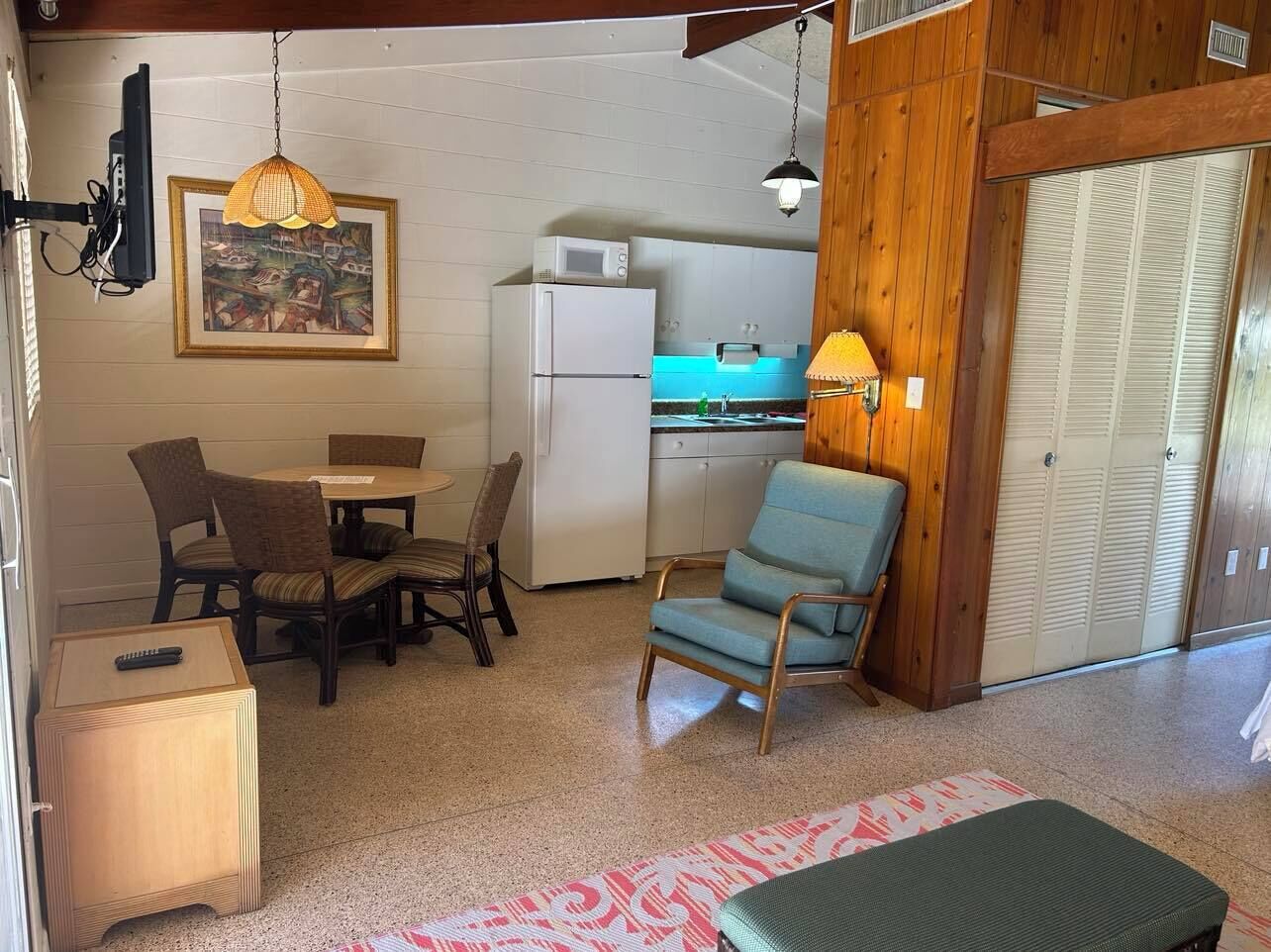 A compact kitchen with a refrigerator, microwave, and wooden cabinets. There is a dining area with a table and chairs, a hanging pendant light, and a framed picture on the wall.