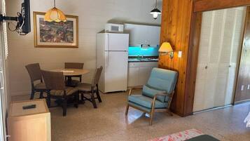 A compact kitchen with a refrigerator, microwave, and wooden cabinets. There is a dining area with a table and chairs, a hanging pendant light, and a framed picture on the wall.