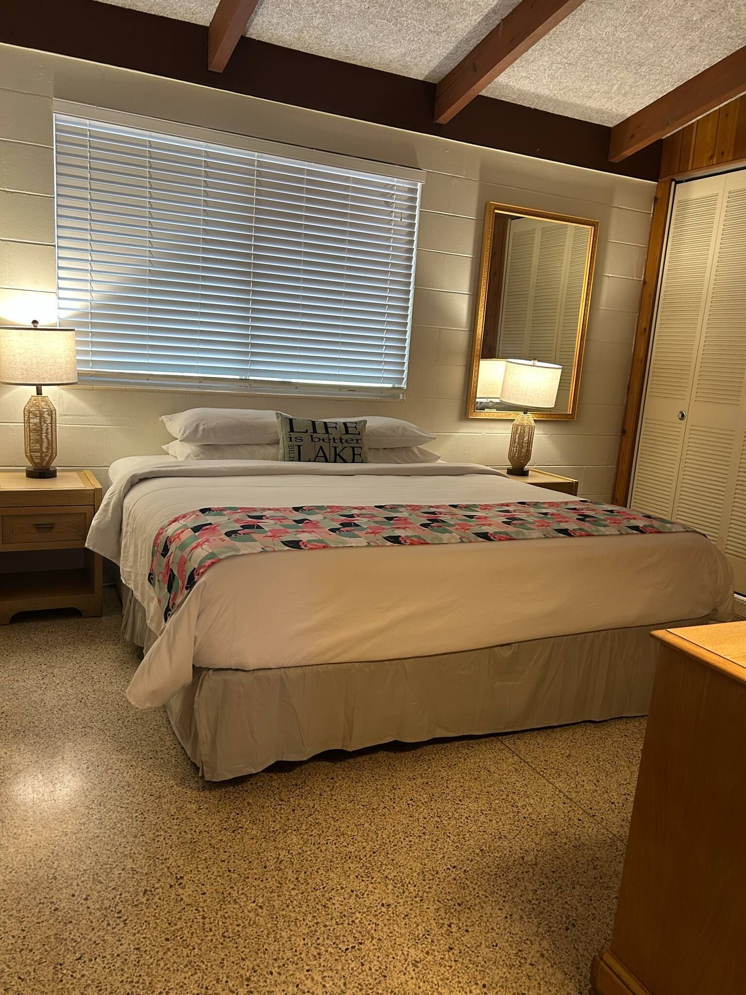 Quarto com cama grande, duas mesas de cabeceira, janela com persianas, espelho e guarda-roupa.