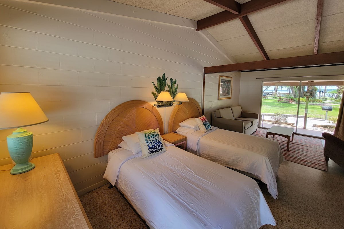 A hotel room with two beds, a wooden headboard, a small table with a lamp, and a view of the outdoors through large windows.