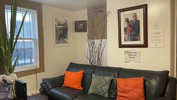A room with a black leather sofa, two orange cushions, a framed photograph, and a small table with a computer and a book.