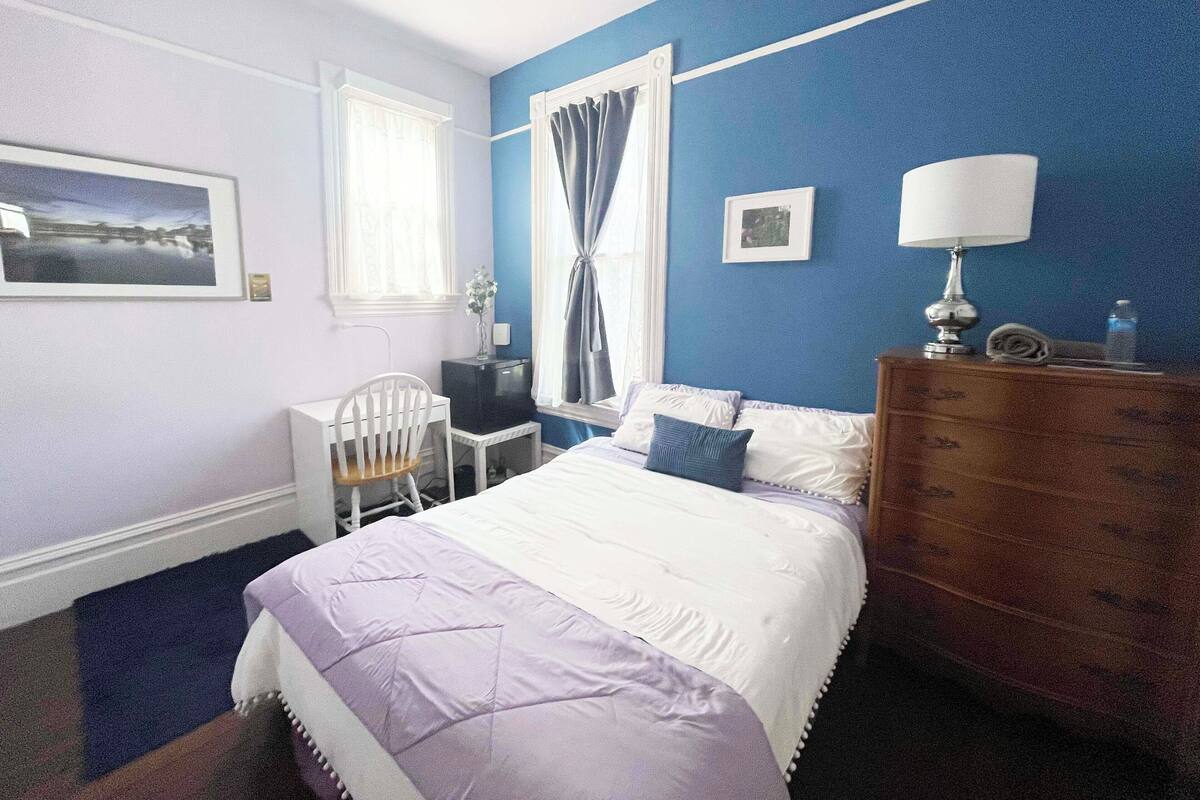 Une chambre à coucher comprenant un lit, un bureau, une chaise, une commode et une fenêtre avec des rideaux.