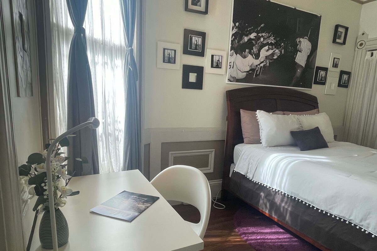 A bedroom with a bed, a desk, a chair, a vase with flowers, and a wall with framed pictures.